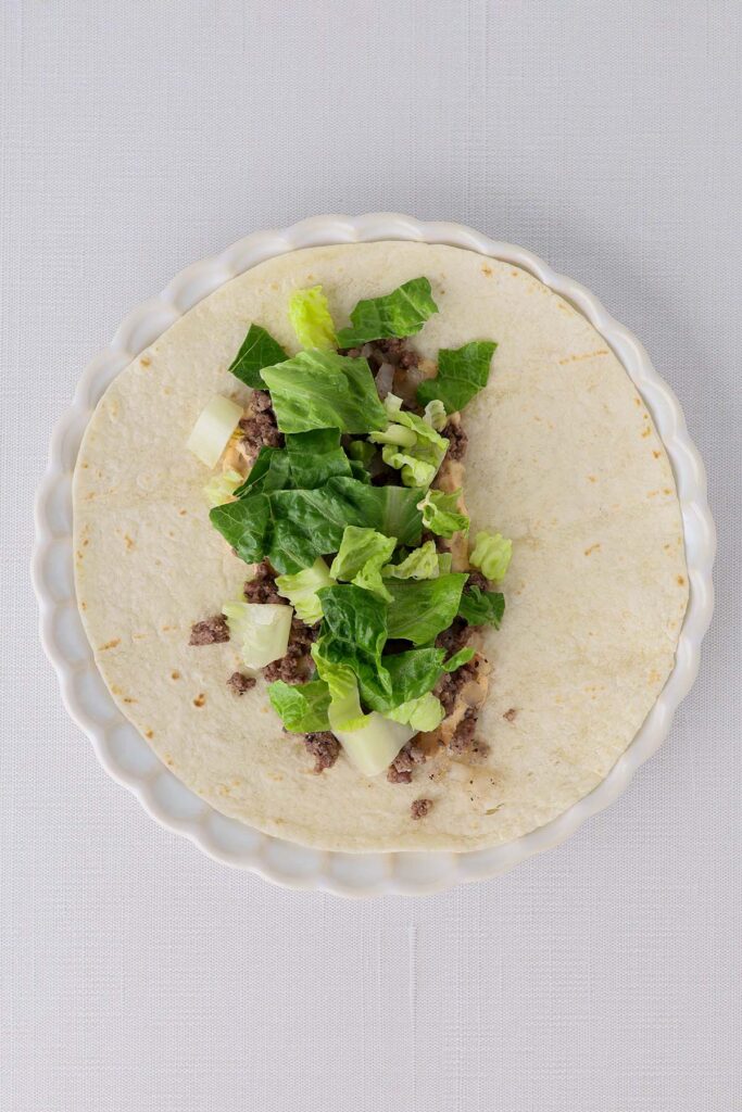 Top down image showing a tortilla on a white plate with burger sauce spread and topped with cooked beef with lettuce