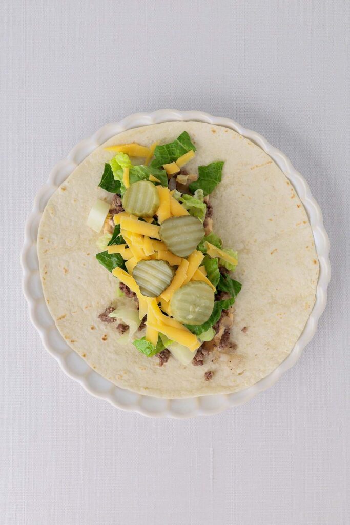 Top down image showing a tortilla on a white plate with burger sauce spread and topped with cooked beef with lettuce cheese and pickles