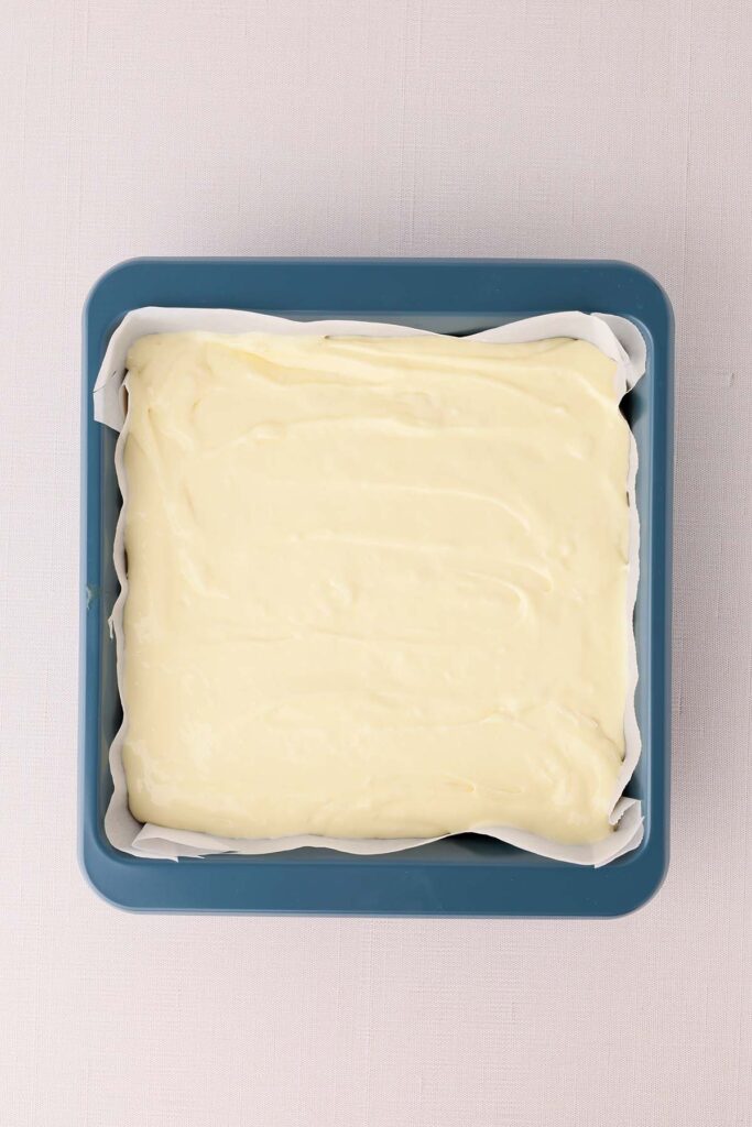 Overhead view of cheesecake filling spread in a parchment-lined square baking pan before baking.