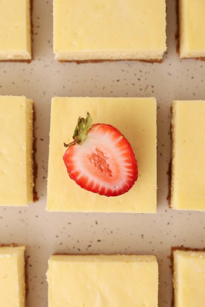 Overhead view of square cheesecake bars arranged in rows on a speckled surface, with one bar topped with a sliced strawberry.