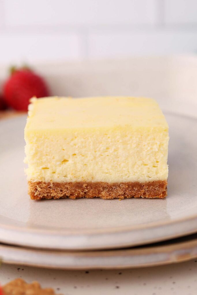 Square cheesecake bar with a graham cracker crust served on a gray plate, showing the thick, creamy cheesecake filling.