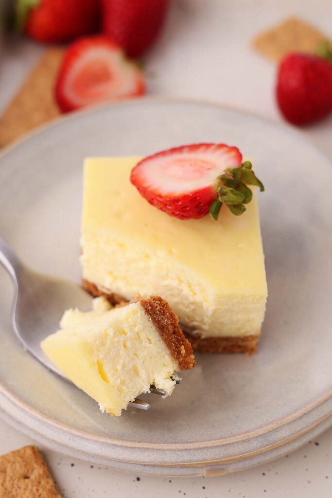 Cheesecake bar with a graham cracker crust topped with a sliced strawberry on a gray plate, with a fork holding a bite of cheesecake in the foreground.