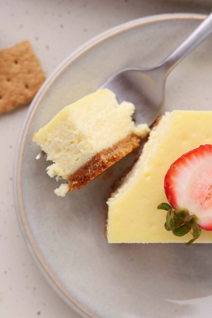 Close-up of a cheesecake bar with a graham cracker crust on a gray plate. A fork holds a bite-sized piece of cheesecake above the plate, and half of a sliced strawberry rests on top of the remaining bar.
