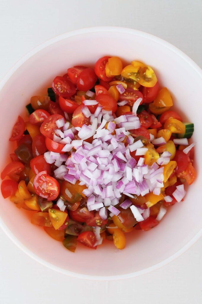 close up top down image showing chopped cucumbers in a bowl with tomatoes on top sprinkled with onion