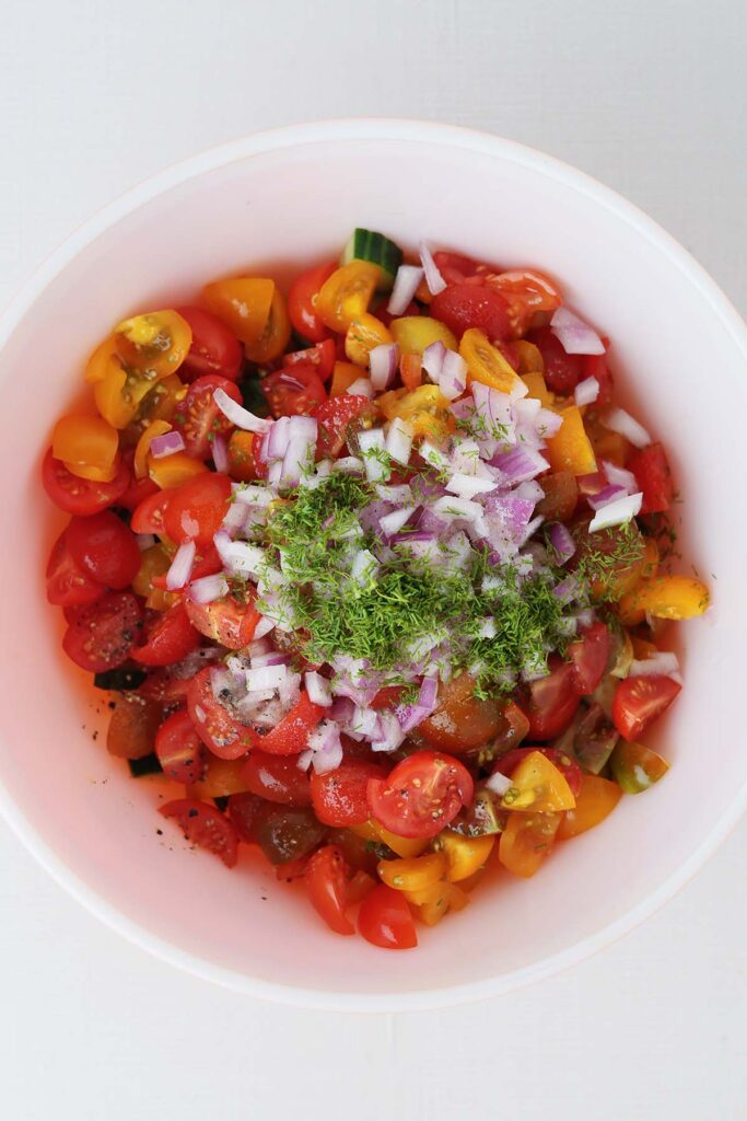 close up top down image showing chopped cucumbers in a bowl with tomatoes on top sprinkled with onion and dill
