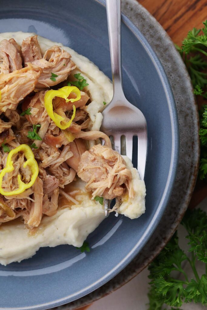 close up image showing a bed of mashed potatoes with shredded chicken, sauce, and yellow peppers on a blue plate. A fork has a bite on it