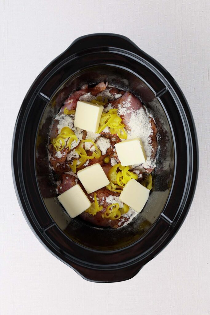 Top down image showing the inside of a black slow cooker insert. It's filled with chicken thighs, seasoning, yellow pepper rings, and butter