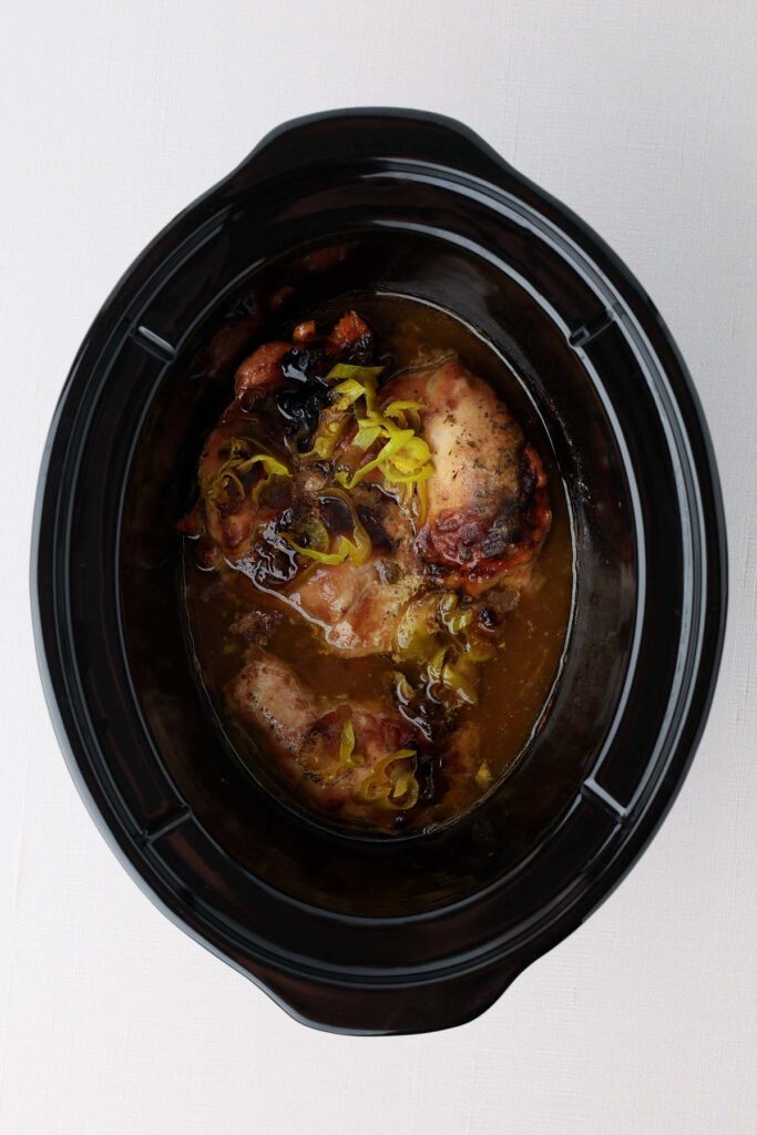 Top down showing the inside of a black slow cooker with cooked chicken, juice, and yellow peppers