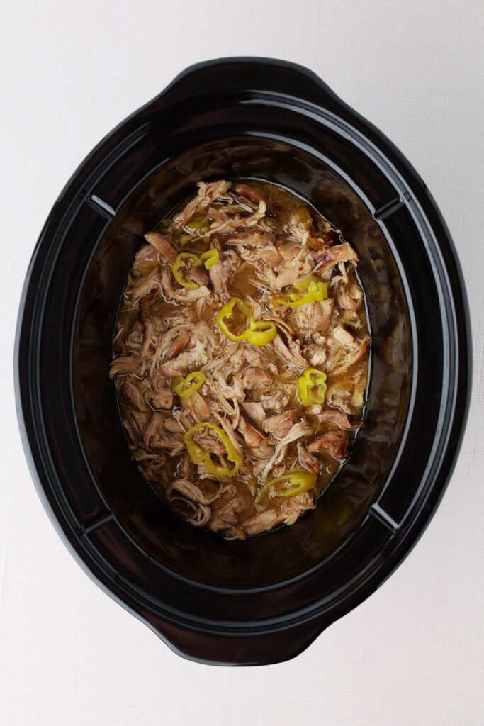 Top down showing the inside of a black slow cooker with cooked shredded chicken, juice, and yellow peppers