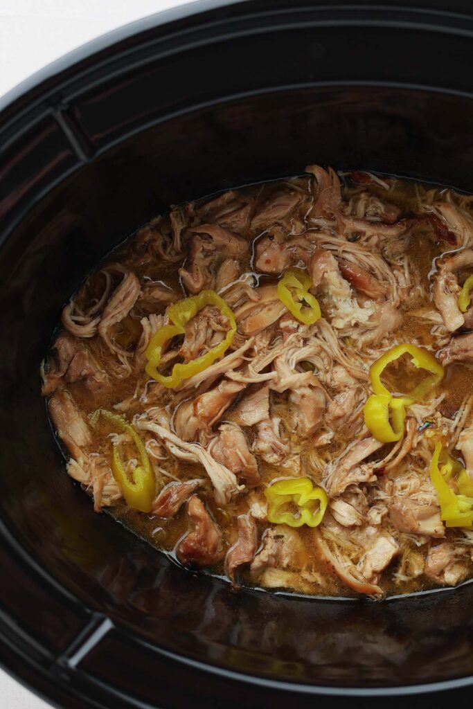 Top down image close into a black slow cooker showing shredded chicken inside liquid with yellow pepper rings on top