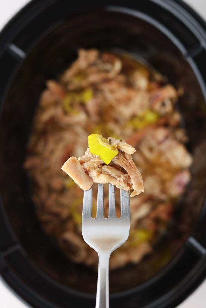 Top down showing the inside of a black slow cooker with cooked shredded chicken, juice, and yellow peppers. A bite is being held over the top with a fork