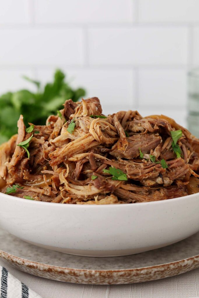 Bowl of shredded pork carnitas topped with chopped cilantro, showing tender strands of slow-cooked pork in cooking juices.