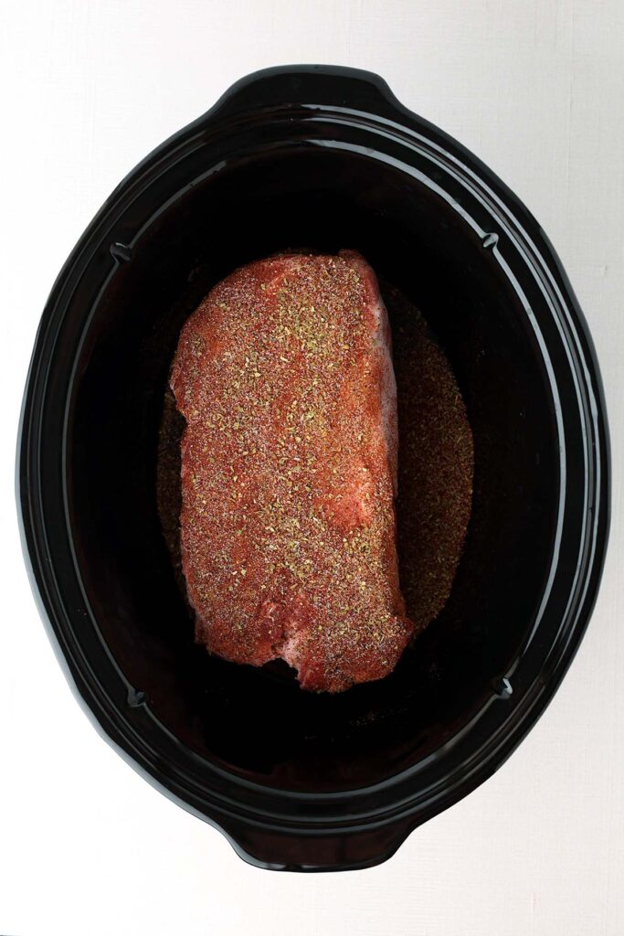 Top down image showing the inside of a slow cooker with a seasoned pork roast