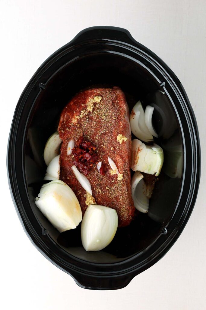 Top down image showing the inside of a slow cooker with a seasoned pork roast, onions, garlic, and chili uncooked
