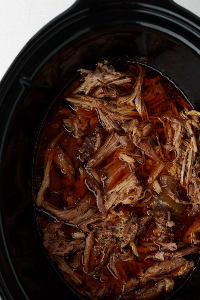Shredded pork carnitas cooking in a slow cooker, surrounded by flavorful cooking juices and softened onions.