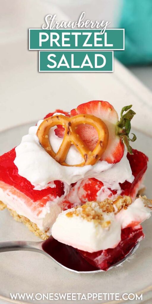 Strawberry pretzel salad served on a white plate with layers of salty pretzel crust, creamy cheesecake filling, and strawberry gelatin topped with whipped cream, a fresh strawberry slice, and a pretzel twist. A spoonful of the dessert is lifted from the plate, showing the distinct layers.