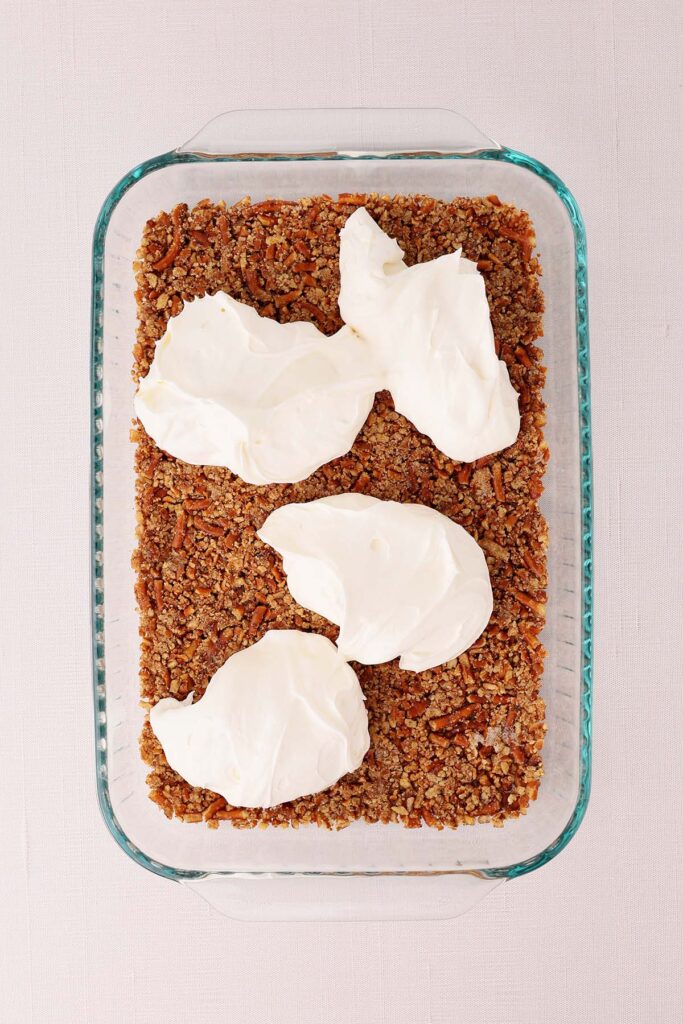 Overhead view of a glass baking dish filled with a buttery crushed pretzel crust topped with dollops of creamy cheesecake filling. This step shows the cream cheese layer being added before spreading it evenly over the pretzel base for strawberry pretzel salad.