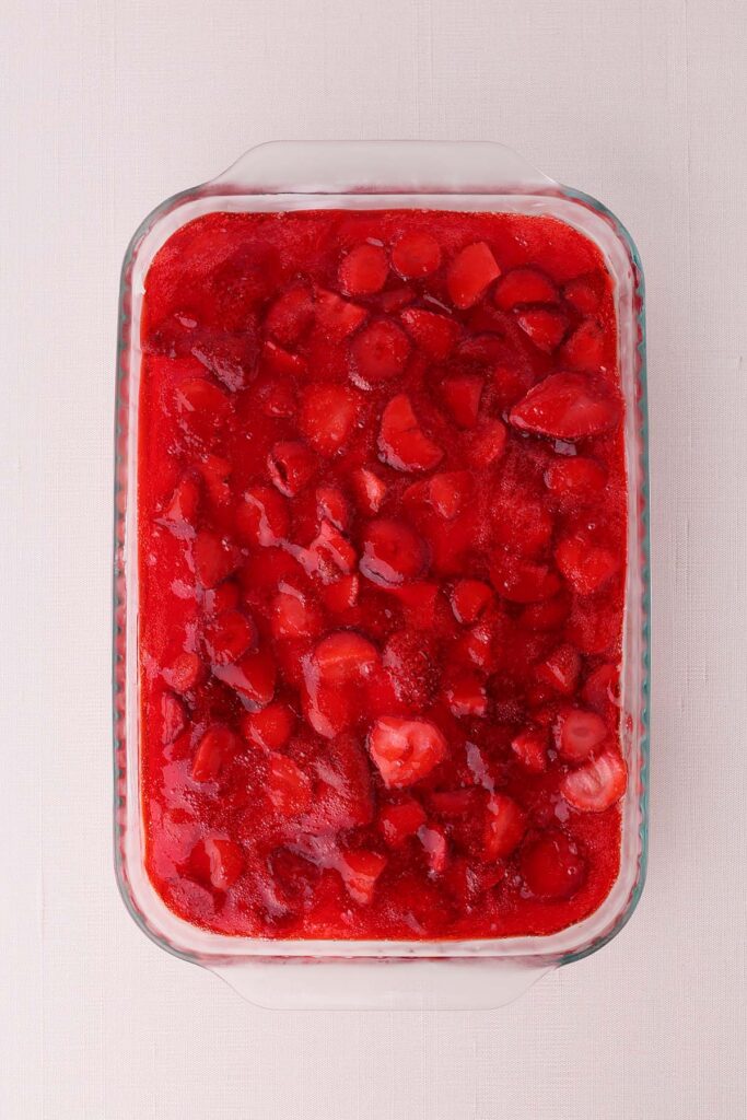 Overhead view of strawberry pretzel salad in a glass baking dish topped with a layer of strawberry gelatin and sliced fresh strawberries. The dessert is shown before serving, highlighting the bright red strawberry topping spread evenly across the pan.