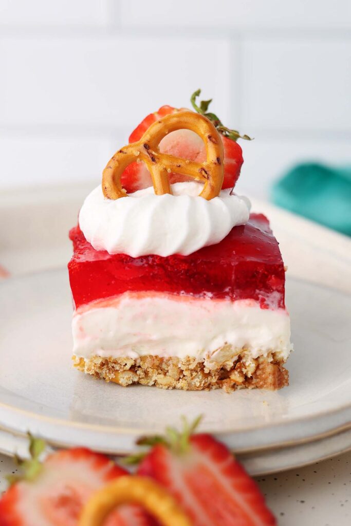 Slice of strawberry pretzel salad on a white plate with layers of buttery pretzel crust, creamy cheesecake filling, and strawberry gelatin. The dessert is topped with whipped cream, a fresh strawberry slice, and a pretzel twist garnish.
