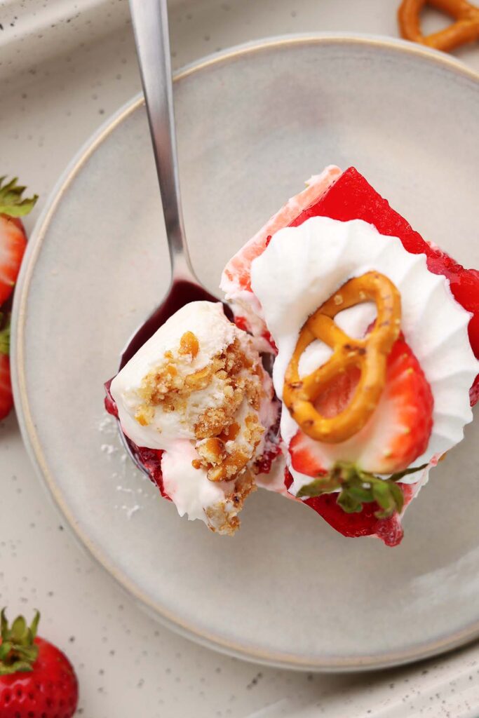 Overhead view of a slice of strawberry pretzel salad on a light plate, featuring layers of pretzel crust, creamy filling, and bright strawberry gelatin. The dessert is topped with whipped cream, a fresh strawberry slice, and a pretzel twist, while a spoon holds a bite showing the creamy filling and crunchy pretzel crust.