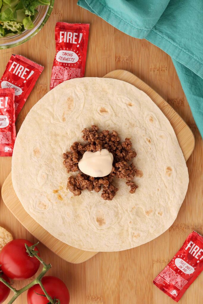 Top down image showing a tortilla with a pile of taco meat in the middle with a dollop of cheese sauce