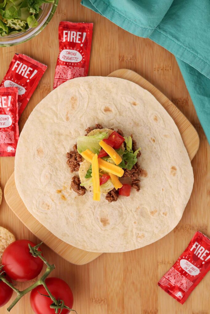 Top down image showing a tortilla with a pile of taco meat in the middle with two tortilla chips, lettuce, tomato, and shredded cheese