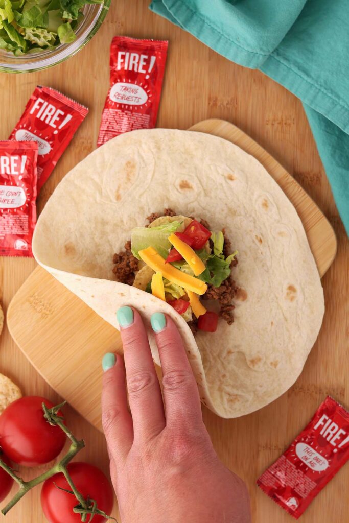 Top down image showing a tortilla with a pile of taco meat in the middle with two tortilla chips, lettuce, tomato, and shredded cheese being folded with a hand