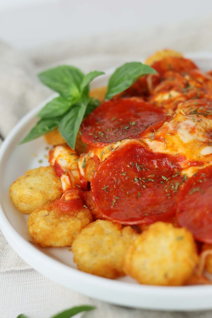 close up image showing a white round plate with a lifted edge topped with crispy tater crowns, sauce, melted mozzarella, pepperoni, and basil