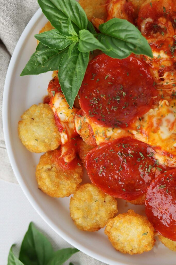 close up image showing a white round plate with a lifted edge topped with crispy tater crowns, sauce, melted mozzarella, pepperoni, and basil