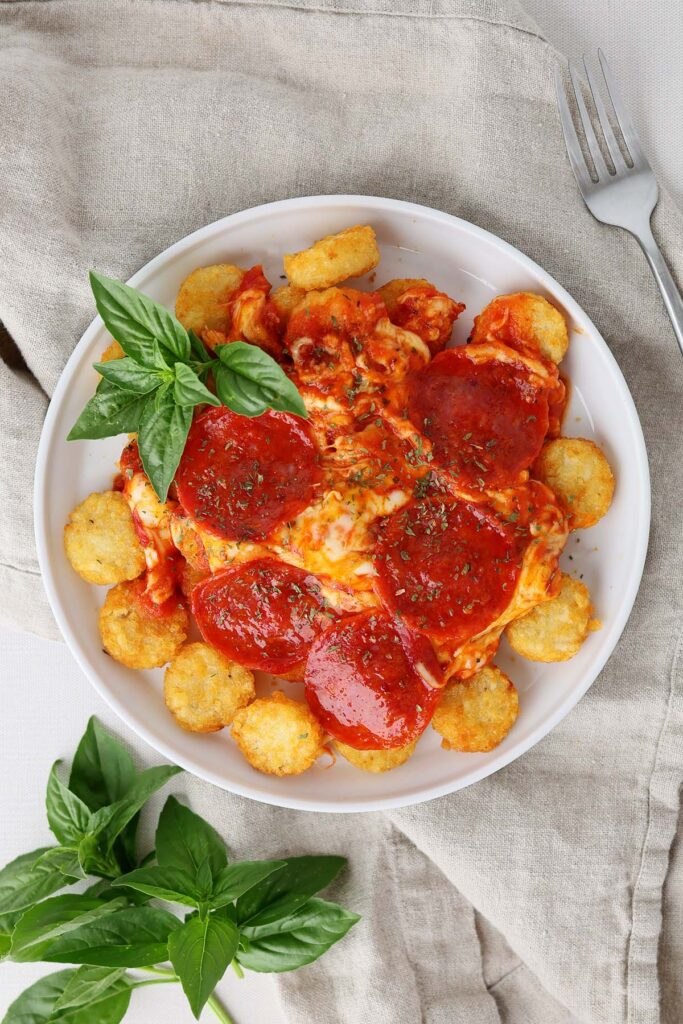 close up image showing a white round plate with a lifted edge topped with crispy tater crowns, sauce, melted mozzarella, pepperoni, and basil