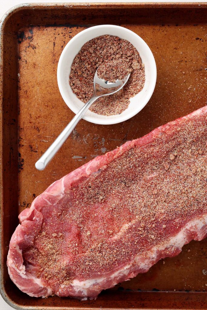 Top down image showing a baking tray with a rack of ribs on top and seasoning next to them in a white bowl with a spoon