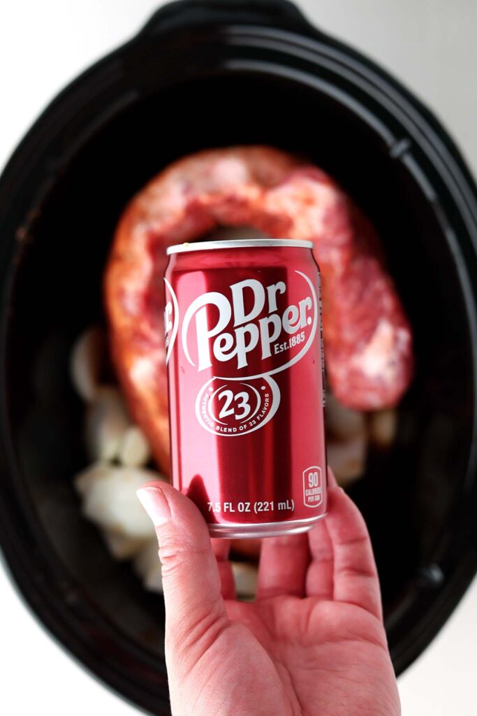Top down image shows a hand holding a small can of dr pepper over a slow cooker with ribs inside