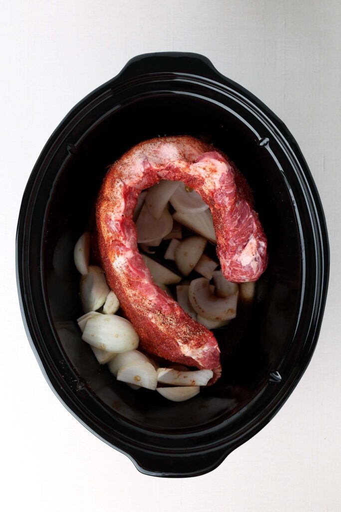 Top down image showing a rack of ribs inside a slow cooker with chopped onion. The ribs are uncooked