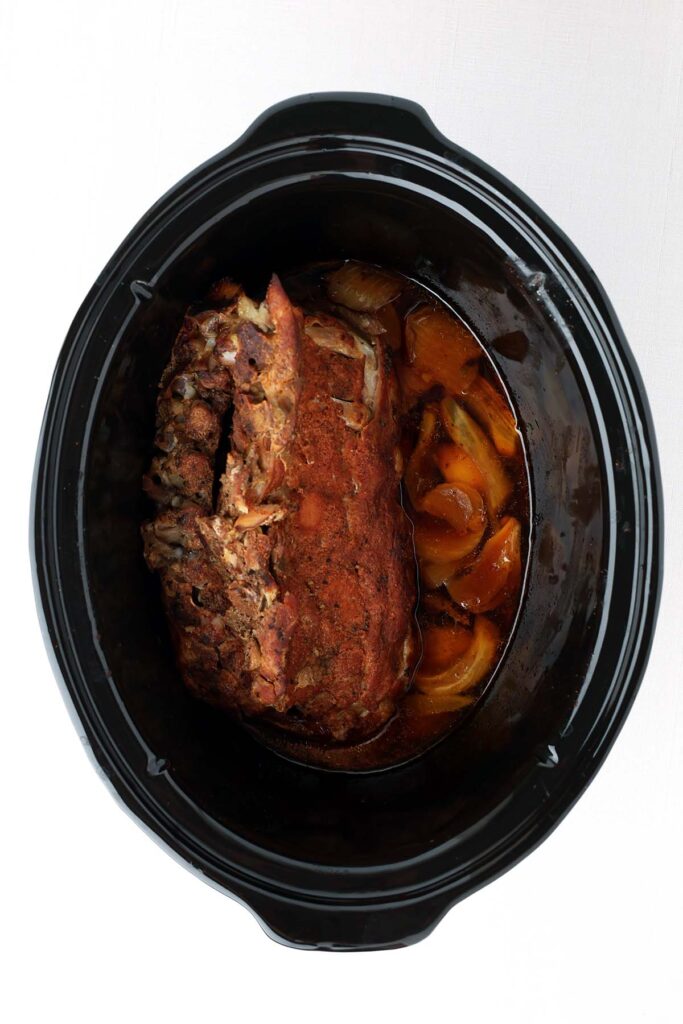 Top down image showing a rack of cooked ribs inside a slow cooker with chopped onion
