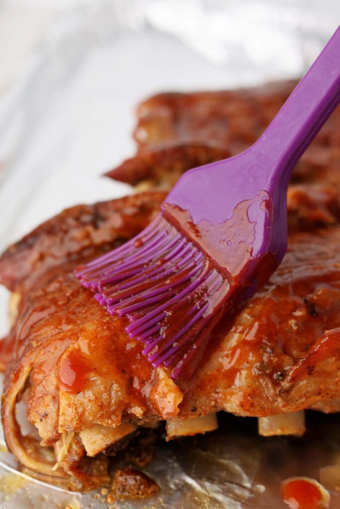 close up image showing a purple silicone brush brushing bbq sauce onto ribs