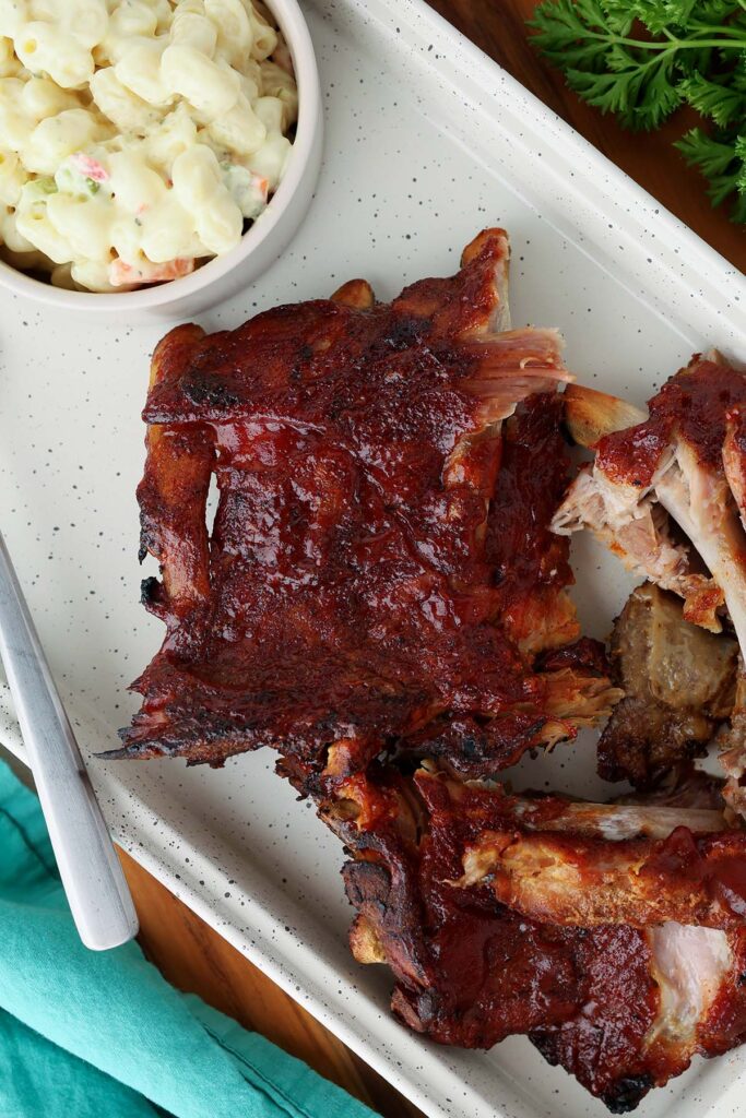 Top down image showing a white tray with ribs scattered across. There's a small bowl of macaroni salad, a fork, and some bbq sauce in a dish on the side
