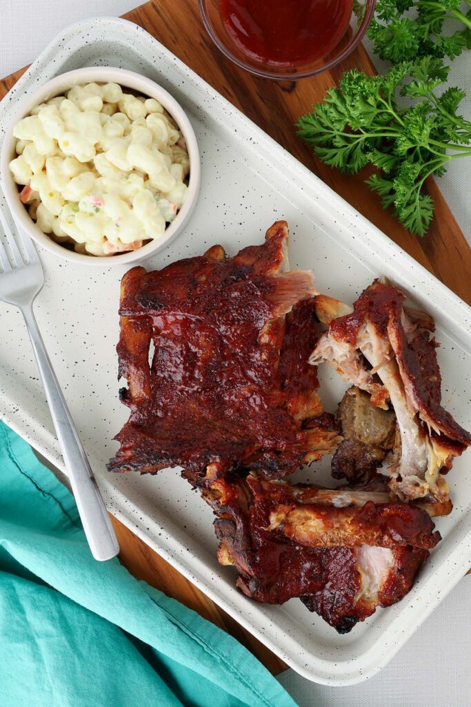 Top down image showing a white tray with ribs scattered across. There's a small bowl of macaroni salad, a fork, and some bbq sauce in a dish on the side