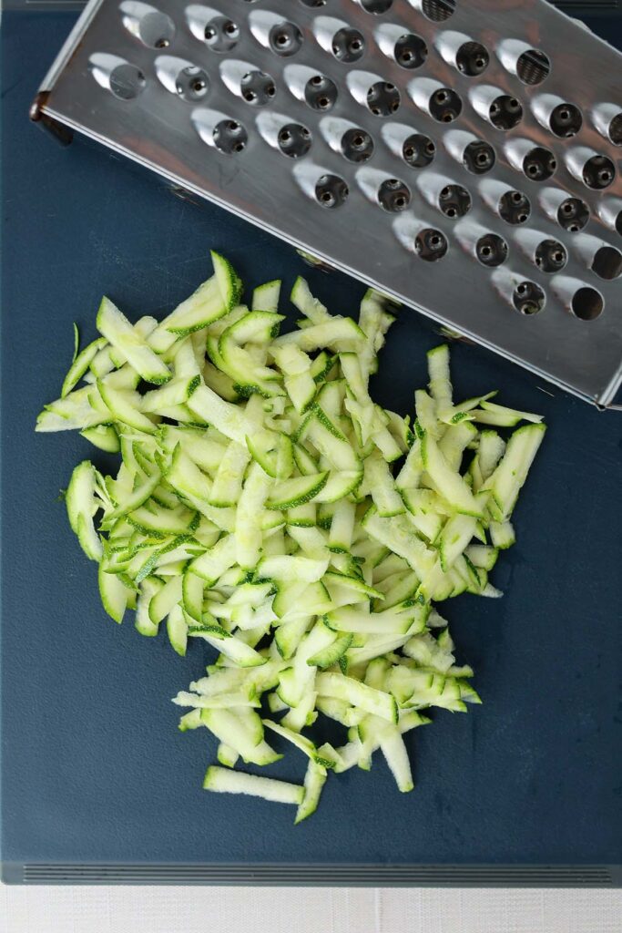 Top down image showing a blue cutting board topped with shreddes of zucchini and a box shredder
