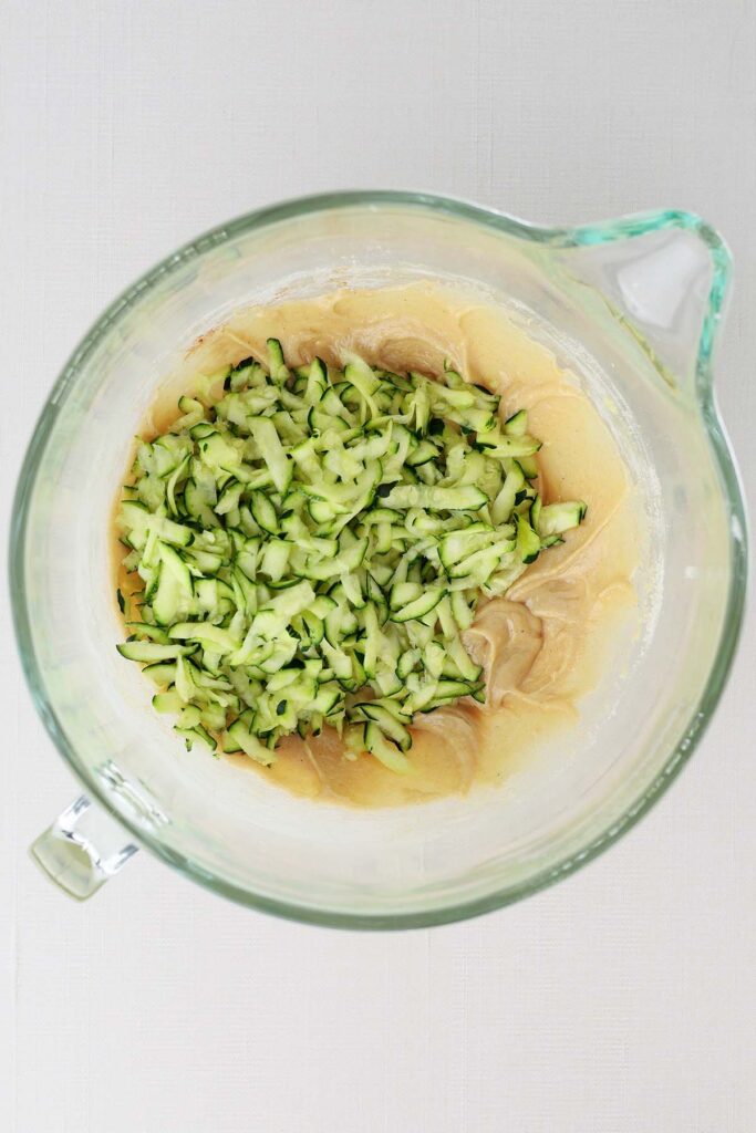 Top down image showing a glass mixing bowl with white batter and shredded zucchini on top