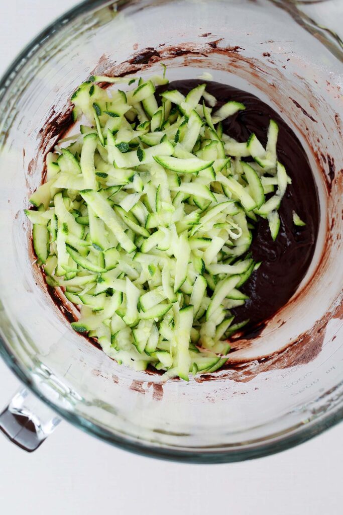 Top down image showing a glass mixing bowl with a chocolate batter and shredded zucchini