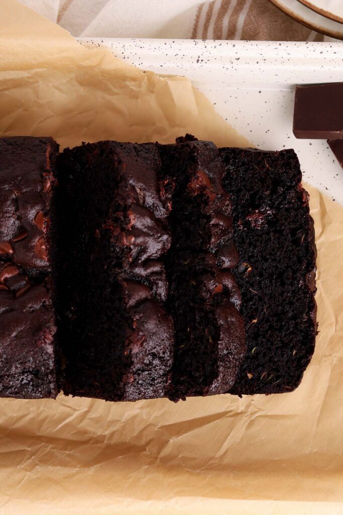 Top down image showing a close up of three sliced chocolate loaf pieces sitting on brown parchment paper on a white tray with pieces of chocolate next to it