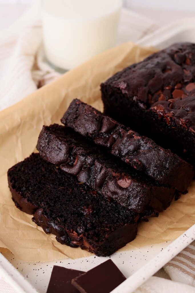 Close up image showing three sliced of chocolate zucchini bread sitting on a brown parchment paper on a white tray with pieces of chocolate next to it.