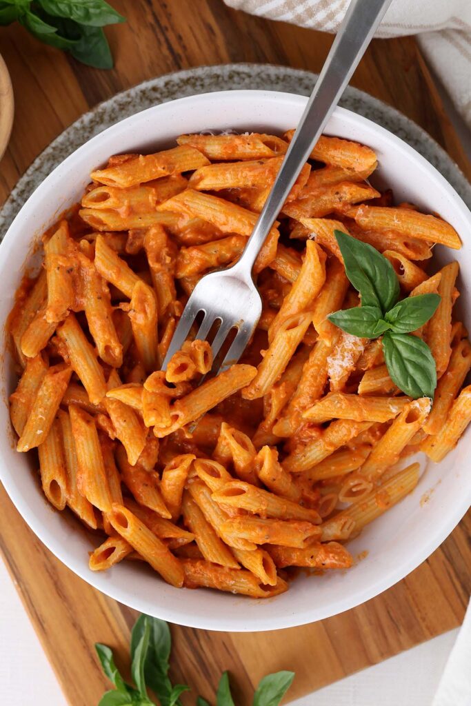 top down image showing a white bowl filled with penne pasta that is coated in a red sauce. There is fresh basil and a bite on a fork