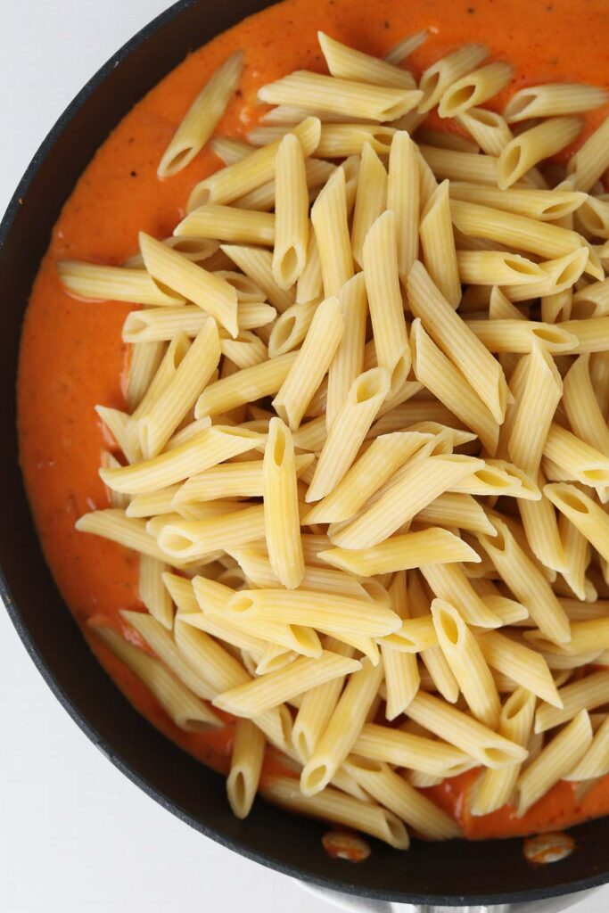 close up top down image showing the edge of a black skillet with a light red sauce with penne sitting on top