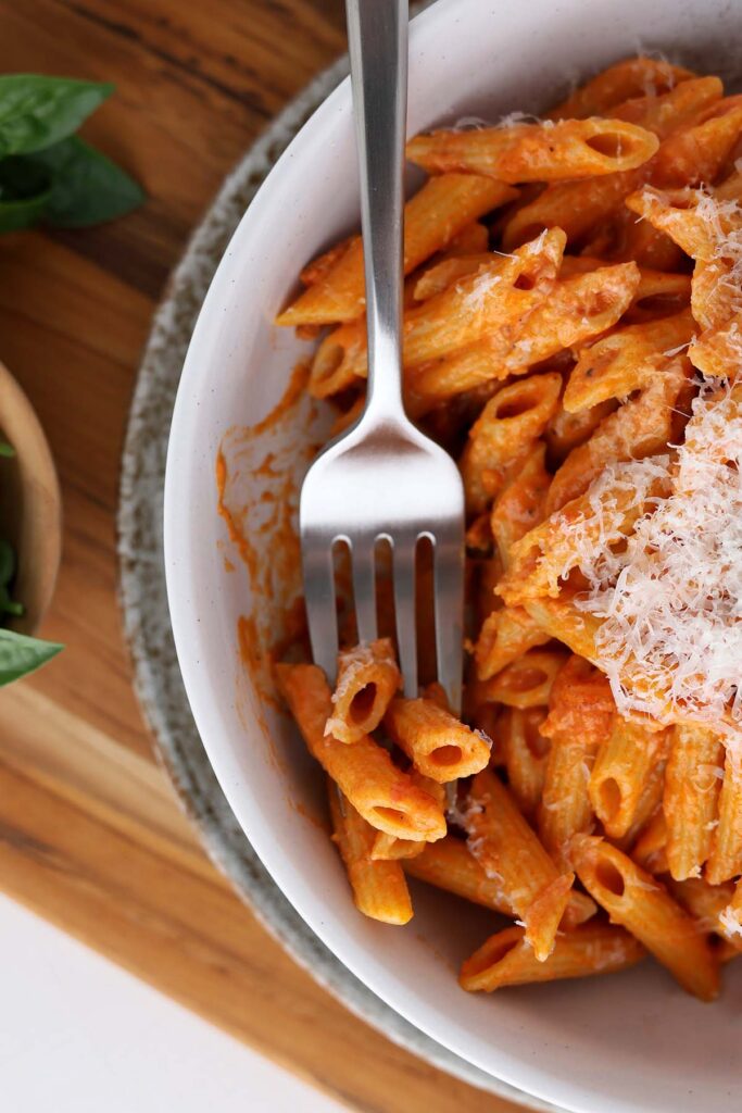 top down imgae showing a close up of a white bowl filled with penne pasta in a red sauce with parmesan cheese, a bite on a fork