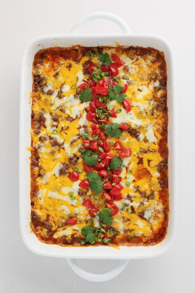 Top down image showing a close up of a white baking dish with red sauce, melty cheese, chopped tomatoes and cilantro