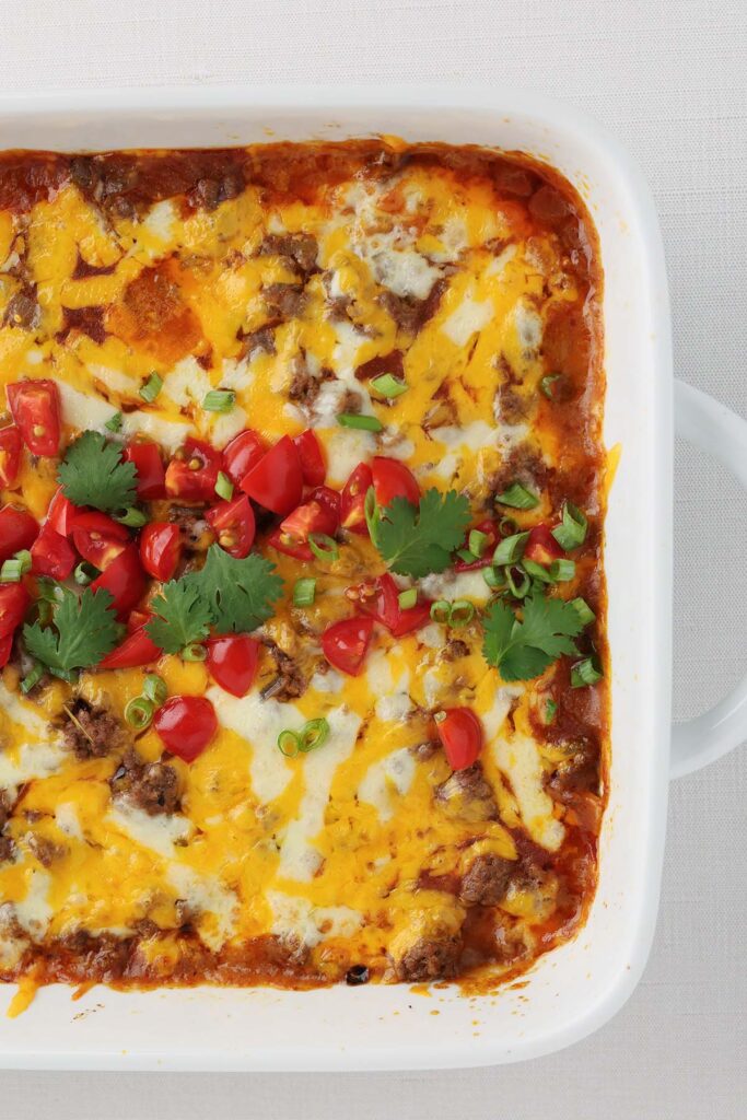 Top down image showing a close up of a white baking dish with red sauce, melty cheese, chopped tomatoes and cilantro