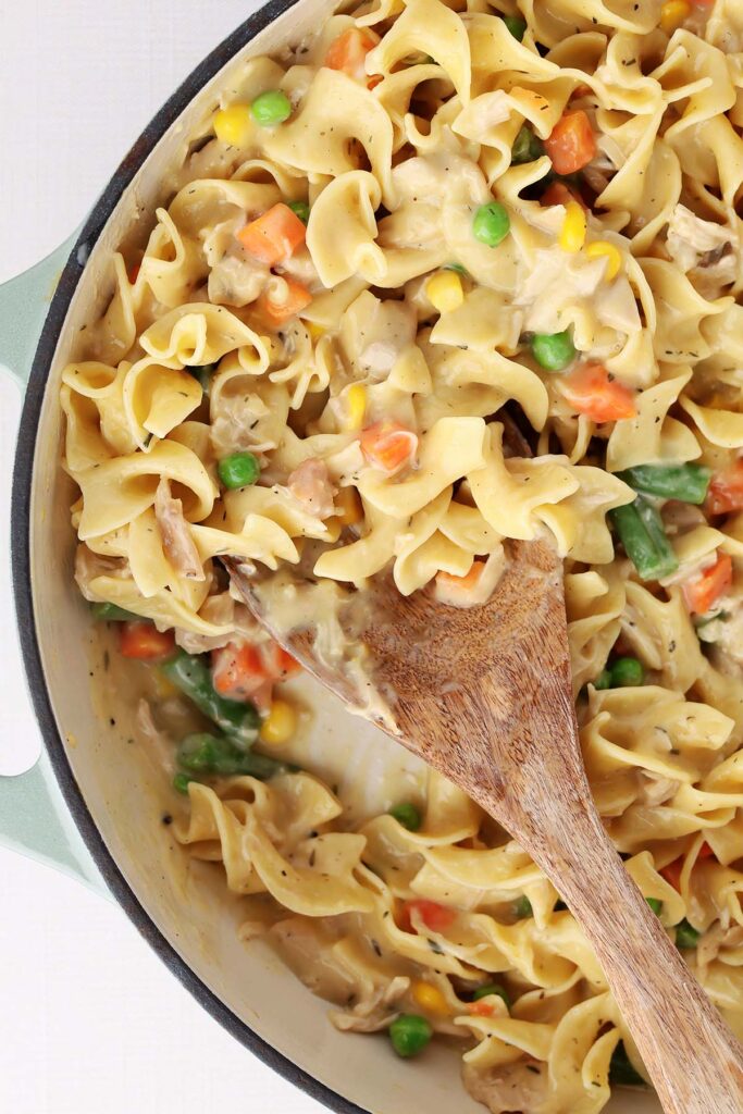 Close up shot of a pot of noodles in a creamy sauce with chicken and mixed veggies with a wooden spoon inside