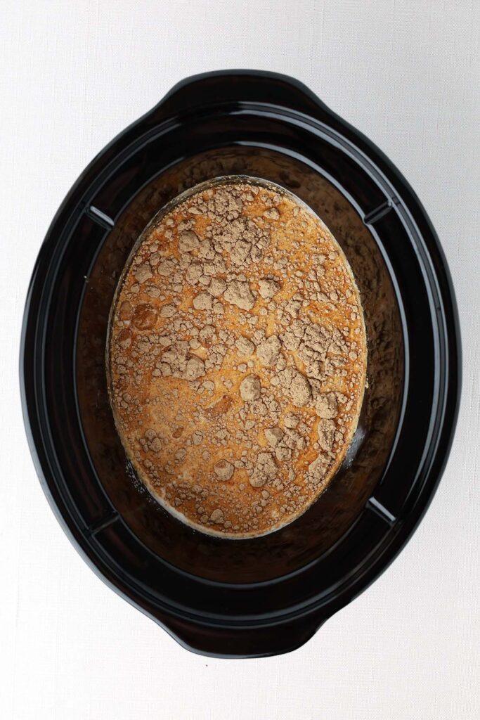 Top down image showing the inside of a slow cooker with liquid and sprinkled with seasoning