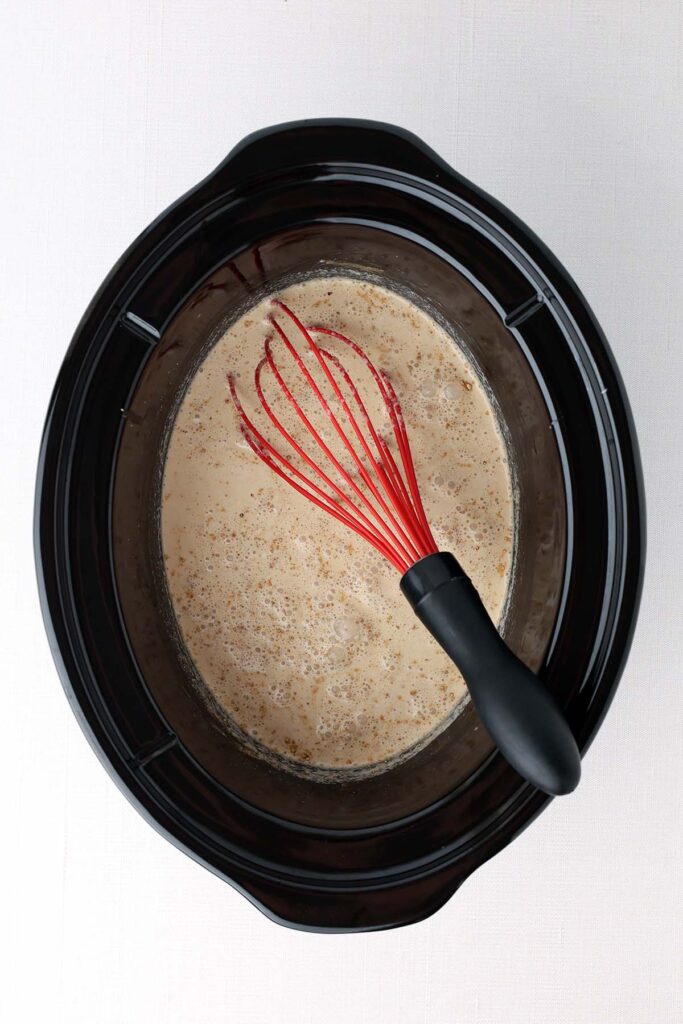 top down image showing the inside of a slow cooker with liquid and a red whisk